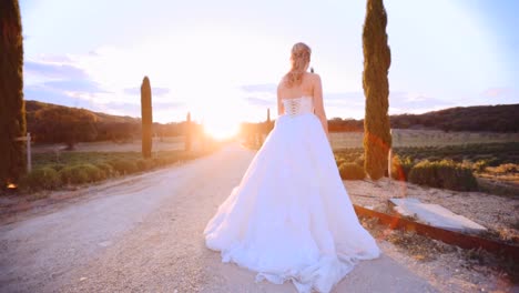 novia en un gran vestido blanco de pie en un camino de macadán en verano al atardecer