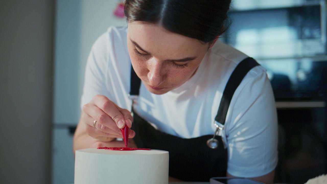 Una donna concentrata decora con cura una torta bianca con glassa rossa. Si china, mostrando precisione e calma concentrazione. Il suo grembiule nero e i capelli ordinati suggeriscono un ambiente professionale.