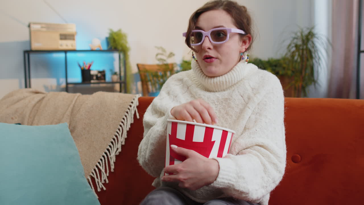 Woman sitting on couch eating popcorn and watching interesting tv 3d film sport game online at home