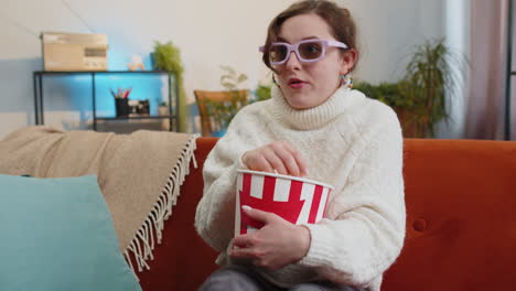 woman sitting on couch eating popcorn and watching interesting tv 3d film sport game online at home
