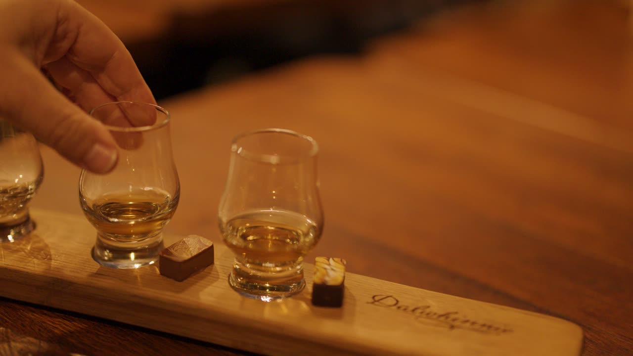 Hand selects whiskey glass from tasting flight with chocolate on wooden tray in warm bar