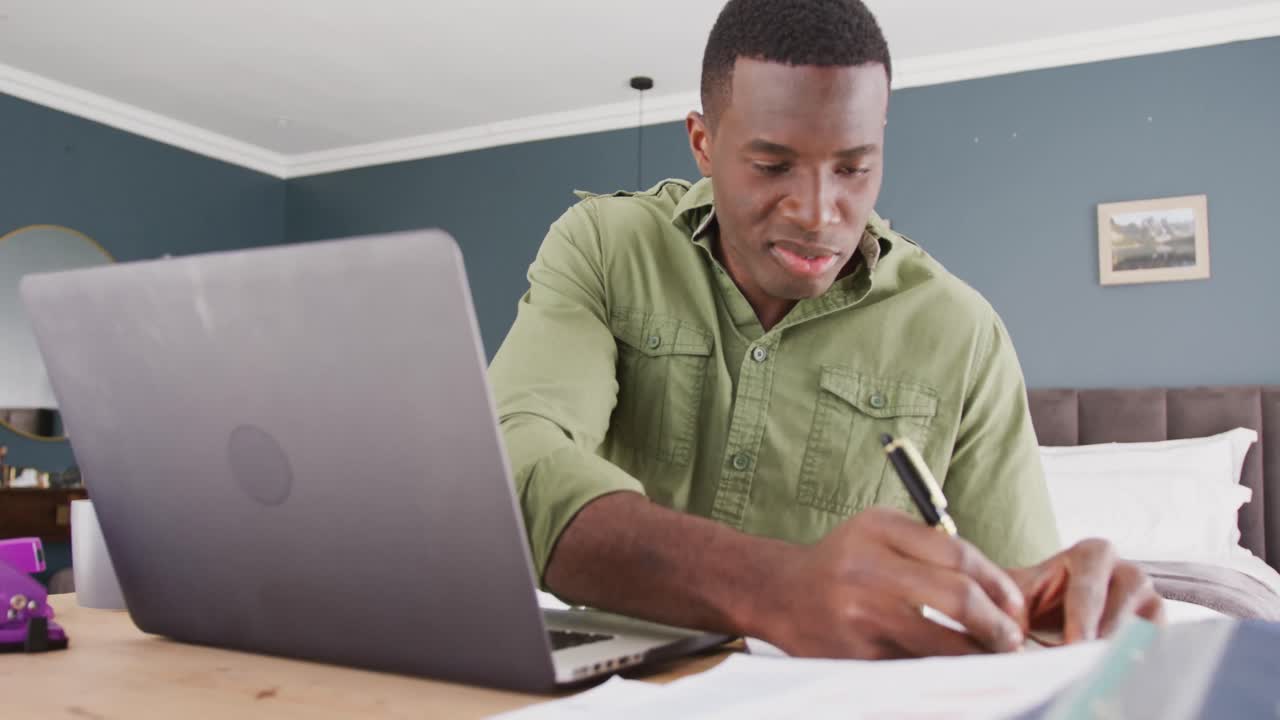 Video of african american man sitting using laptop and writing notes, working in bedroom