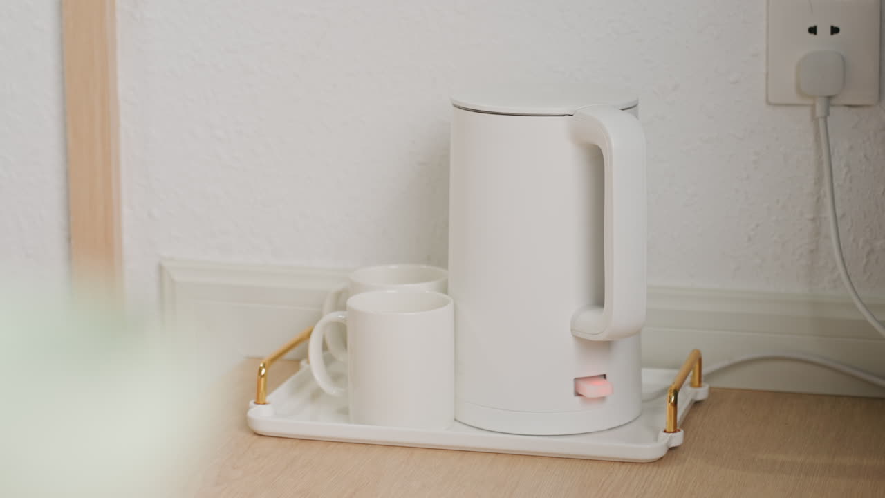 Bollitore elettrico pronto sul vassoio dell'hotel, elettrodomestico compatto bianco con tazza su vassoio decorativo, mano che preme l'interruttore, cavo collegato alla presa a muro, un'ordinata comodità alberghiera per il ristoro dell'ospite.