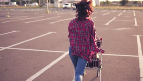 Back-view-of-a-young-woman-sitting-in-the-grocery-cart,-while-her-friend-is-pushing-her-behind-in-the-parking-by-the-shopping
