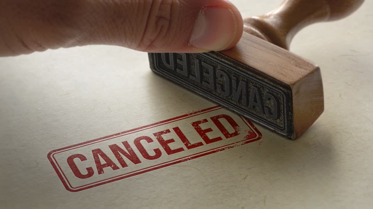 Lowering hand pressing wooden stamp onto textured paper on desk, marking document CANCELED in red