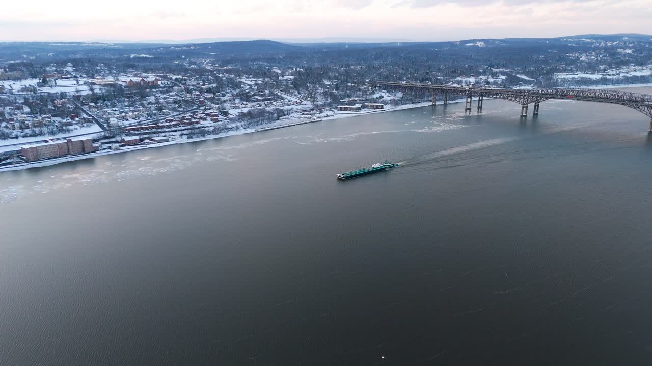 Striking aerial perspective of a cargo ship gliding across the Hudson River, with remnants of ice on the water's surface. The scene features snow-covered hills and a quaint town, highlighting the sere