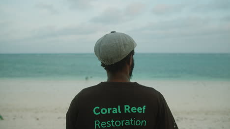 Medium-shot-of-a-young-black-man-with-his-back-to-camera-looking-out-into-the-ocean-wearing-a-coral-reef-restoration-t-shirt