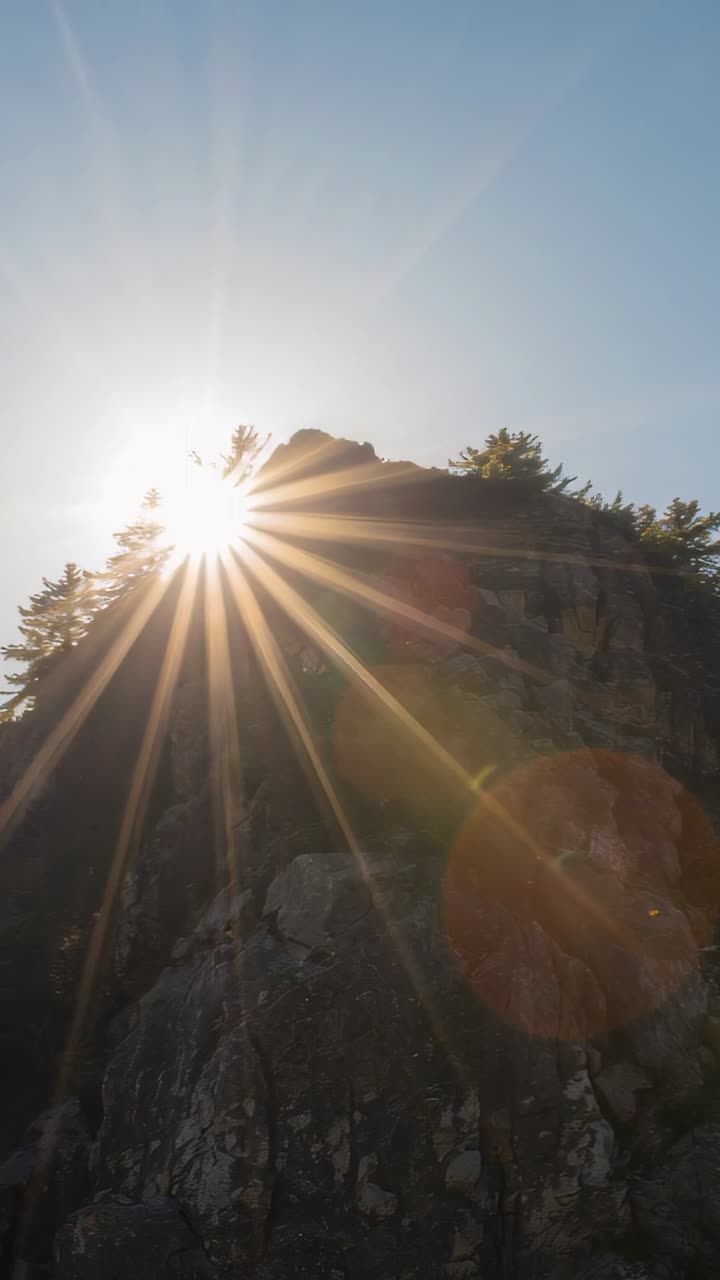 Vertical video: Moving sun sliding behind rock cliff from below, making starburst flares over trees