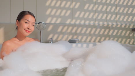 woman taking a hot bath relaxing in a bathtub filled with foam outside under sunlight, asian female looking into camera and smiles