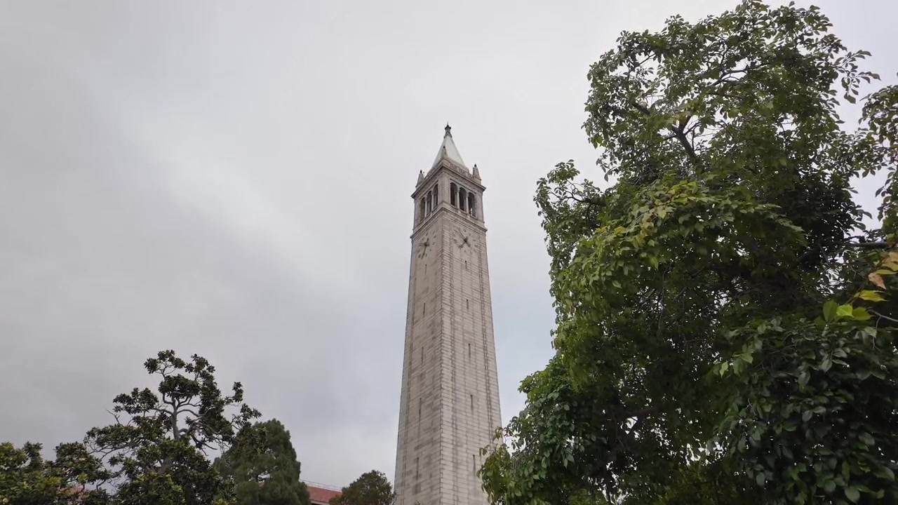 Toma de gran angular de la Torre Sather en UC Berkeley, que se eleva sobre vibrantes árboles verdes contra un cielo nublado, mostrando una arquitectura impactante y una atmósfera tranquila en el campus.