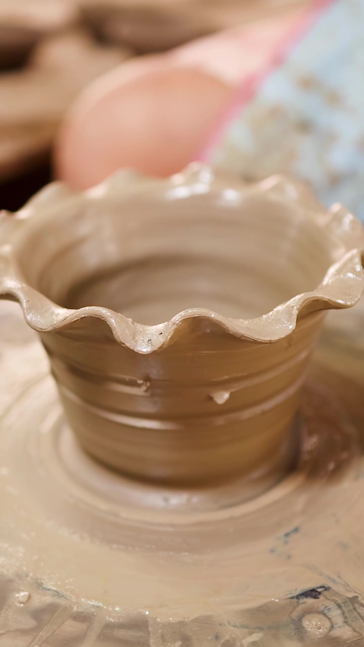A wet terracotta clay pot with scalloped edges rotates on a spinning pottery wheel.