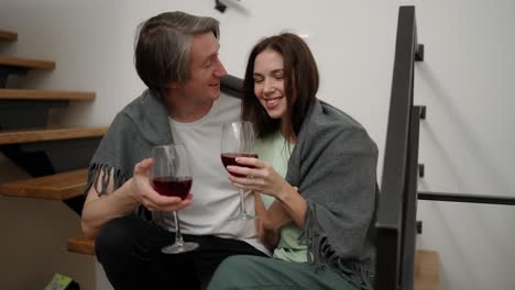 Smiling-couple-sit-on-the-wooden-stairs-hold-glasses-drink-red-wine-talk-laugh-hugging