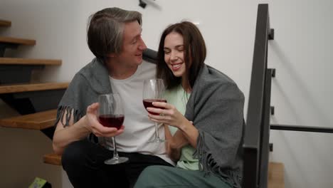 una pareja sonriente sentada en las escaleras de madera sosteniendo copas bebiendo vino tinto hablando riendo abrazándose