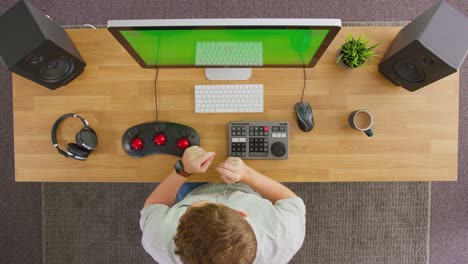 overhead view of male video editor putting on wireless headphones and working at computer with green screen in creative office - shot in slow motion