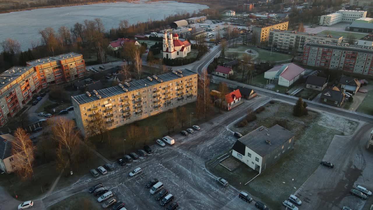 Aerial photo of Balvi city bathed in warm sunset light, featuring urban buildings, parking lots, and a snow-dusted landscape. A peaceful winter scene.