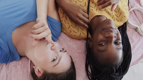 portrait of happy diverse teenage female friends lying on their backs on bed and taking selfie