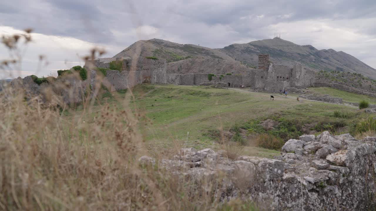 Stone ruins of Rozafa Castle with partial towers and walls surrounded by grassland near Shkodër, Albania. Mountains in the background. Static view