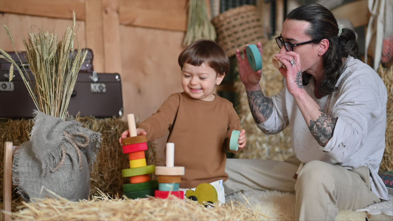 Father playing with his son with colourful, ecological wooden toys in a barn, near square hay bales