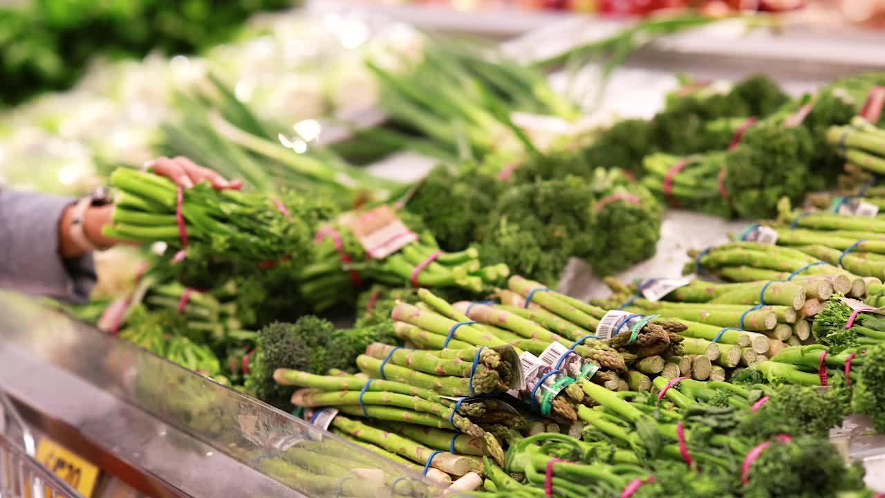 A person picks up broccoli from a well-lit supermarket produce section, surrounded by vibrant green vegetables