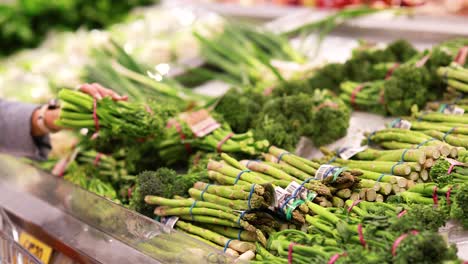a person picks up broccoli from a well-lit supermarket produce section, surrounded by vibrant green vegetables