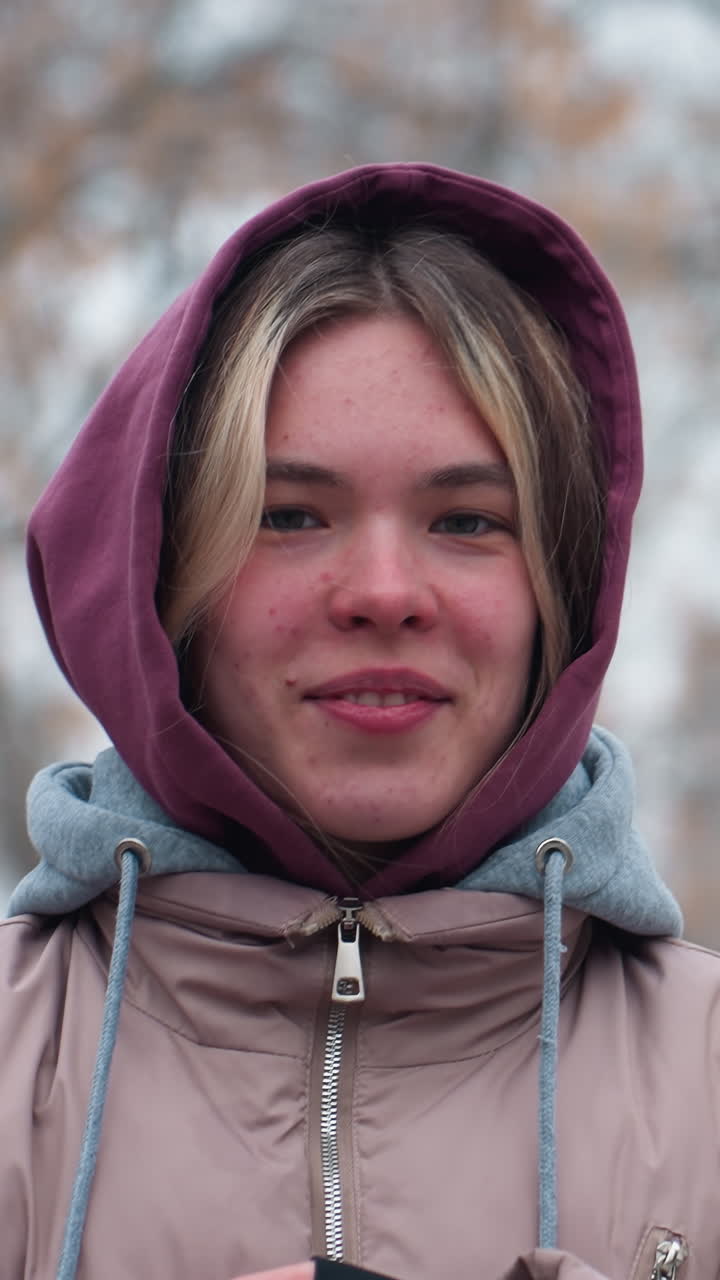 joven con traje de invierno con manchas en la cara sosteniendo una cadena de columpio y apoyando su cabeza en ella, con una chaqueta con capucha, expresión relajada, fondo nevado borroso con árboles desnudos