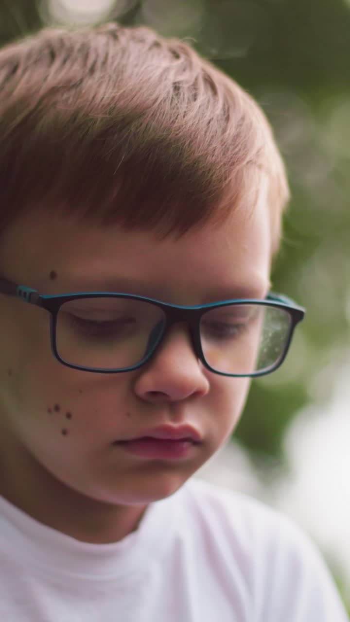 un niño pequeño con gafas parece abatido y pensativo, mirando hacia abajo con un fondo borroso de vegetación y una vista parcial de la cabeza de una mujer