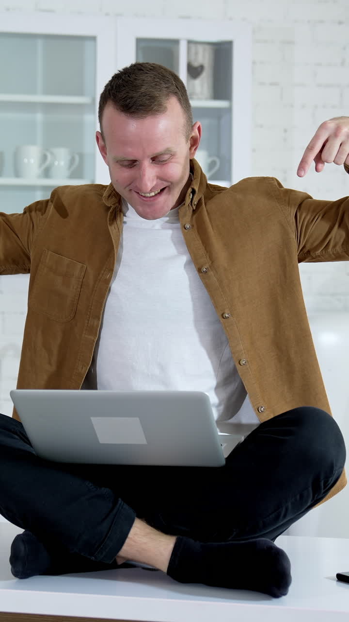 Funny man with a laptop on a table.Smiling freelancer sitting on a table and waiting to press button on a laptop on the kitchen background. Vertical video
