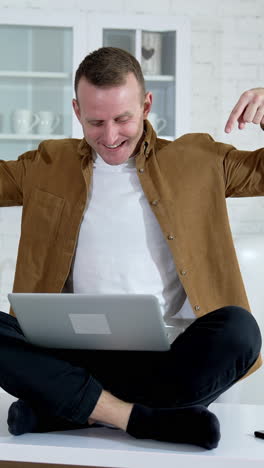 funny man with a laptop on a table.smiling freelancer sitting on a table and waiting to press button on a laptop on the kitchen background. vertical video