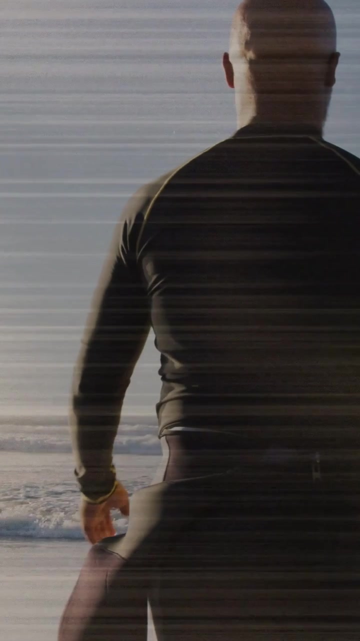 Walking man in dark wetsuit holding green surfboard with wristband toward ocean on beach, to surf