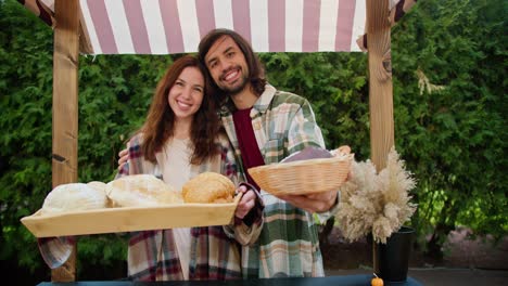 A-happy-brunette-guy-and-his-brunette-girlfriend-in-checkered-shirts-hold-out-trays-with-delicious-pastries-while-working-in-a-shop-against-the-backdrop-of-green-coniferous-trees-in-the-summer