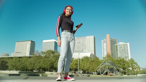 Empowered-Young-Latin-Woman-plays-music-with-the-mobile-using-wireless-red-headphones-and-starts-dancing-on-a-sunny-day-at-the-city