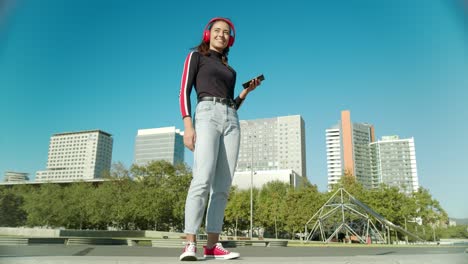 Una-Joven-Latina-Empoderada-Toca-Música-Con-El-Móvil-Usando-Auriculares-Rojos-Inalámbricos-Y-Comienza-A-Bailar-En-Un-Día-Soleado-En-La-Ciudad