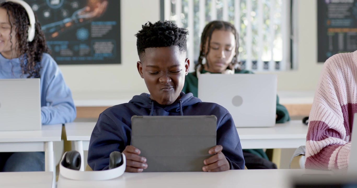 Teen classmates working in class, navy-hoodie teen holding tablet, others typing laptops