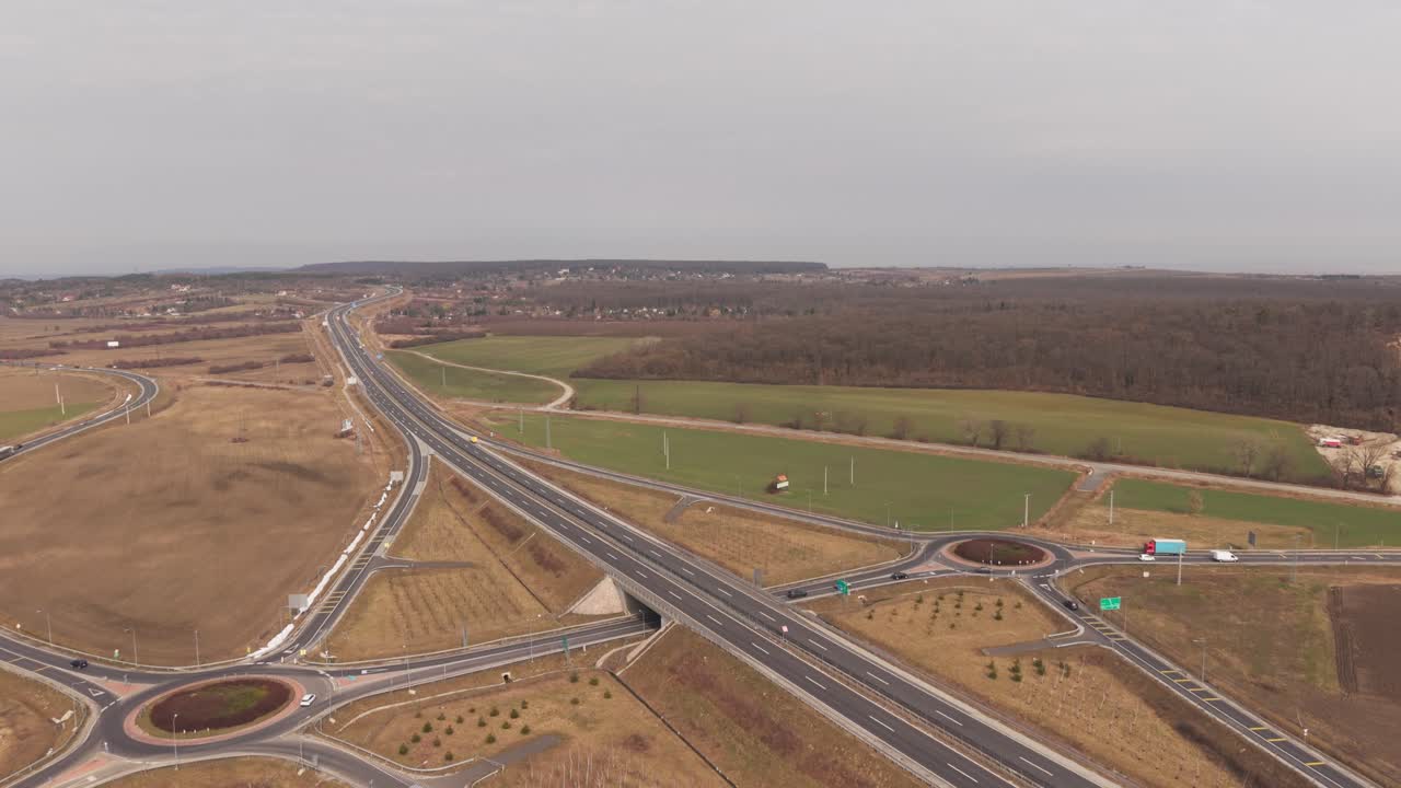 Veduta aerea con drone di un moderno svincolo autostradale con rotatorie che collegano più strade attraverso un paesaggio rurale. Campi agricoli e campagna aperta circondano la rete stradale.