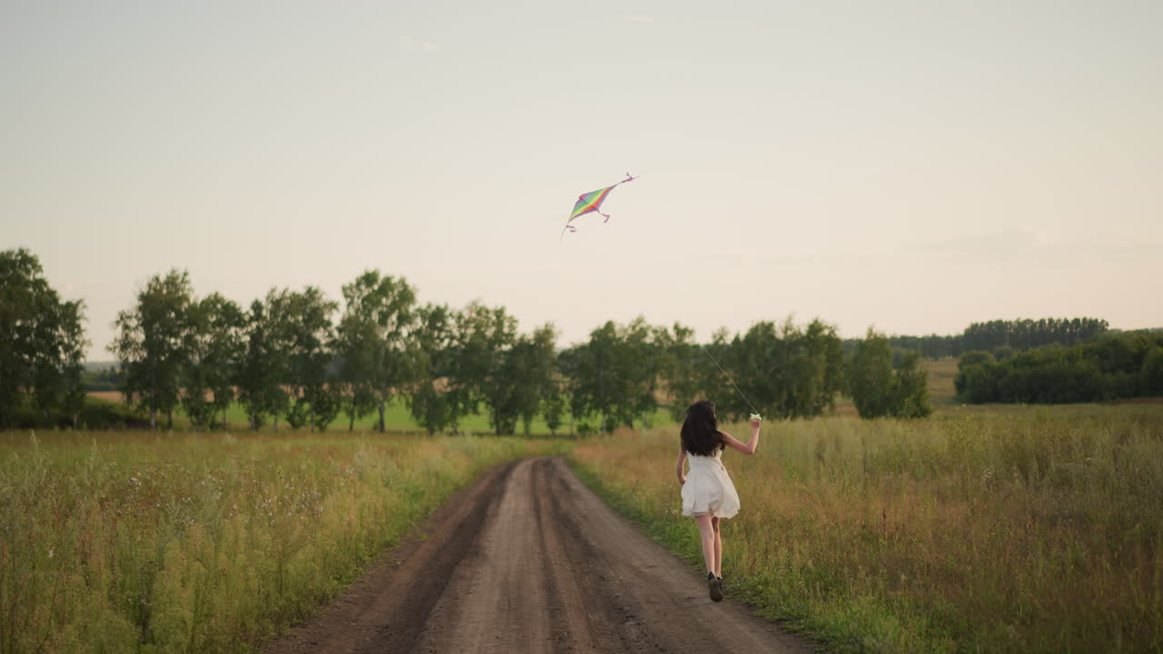 Child pursues flying kite scenic, Young girl chases kite along country lane during twilight atmosphere, Female child follows her kite across rural road at sunset capturing nostalgic mood