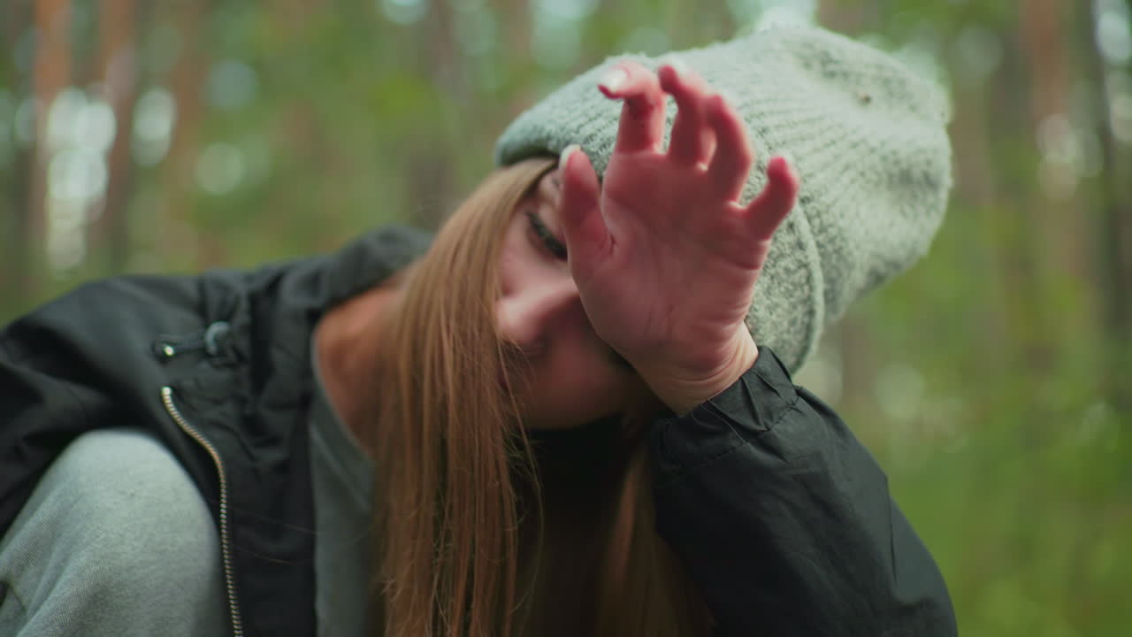 woman in gray beanie and black jacket wipes sweat from forehead with hand while walking in forest, showing tired expression and long hair falling across face in soft natural light