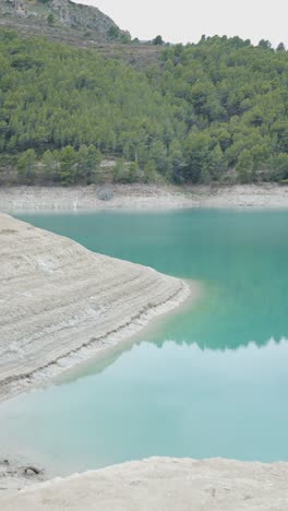 a person photographs a turquoise lake with a dry, sandy shore and green forest