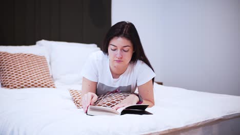 Una-Joven-Está-Leyendo-Un-Libro-Con-Tapa-Dura-Negra-En-Su-Dormitorio,-De-Aspecto-Serio,-Acostada-En-La-Cama,-Pasa-Su-Tiempo-Libre-Con-Un-Libro,-Relajándose-En-Casa,-Pasa-La-Página,-Voltea-La-Página