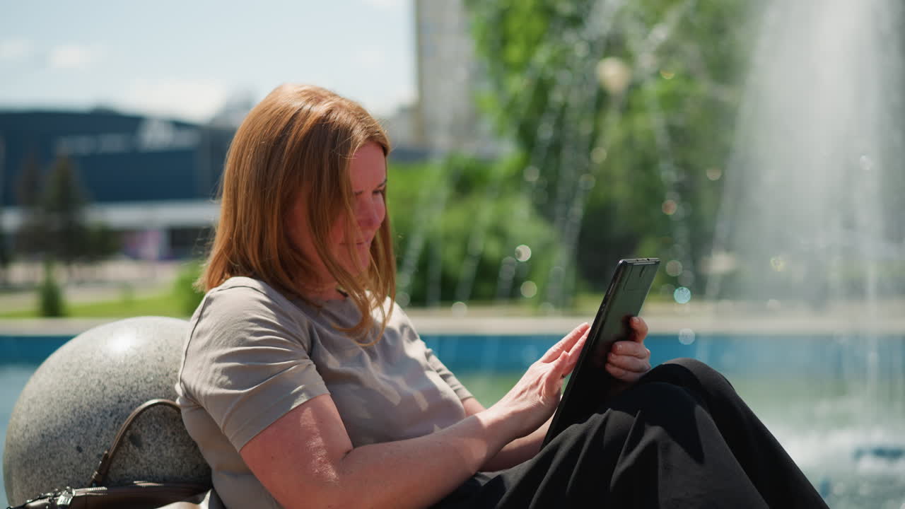Young lady sitting relaxed by city fountain using tablet smiling as she slides finger on screen, brown bag resting beside, surrounded by greenery and sunlight
