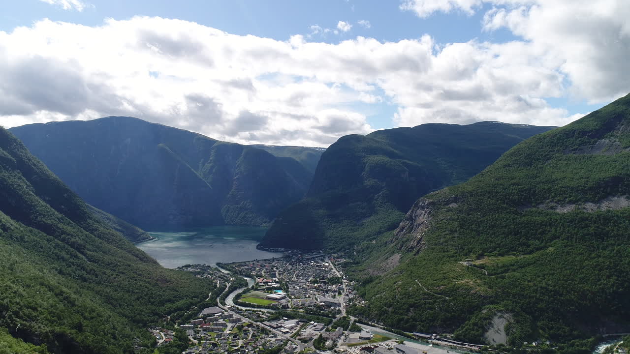 노르웨이 서쪽 해안의 활기찬 마을의 공중 풍경, 부분적으로 구름이 많은 파란 하늘 아래 초록색 산으로 둘러싸인 피오르드 계곡에 자리 잡고 있습니다.