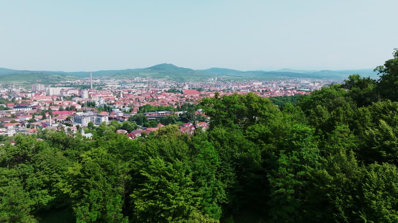 A sweeping aerial wide shot revealing a dense, a vast European cityscape Bistrita of residential and commercial buildings stretching towards distant hills under a bright, clear sky