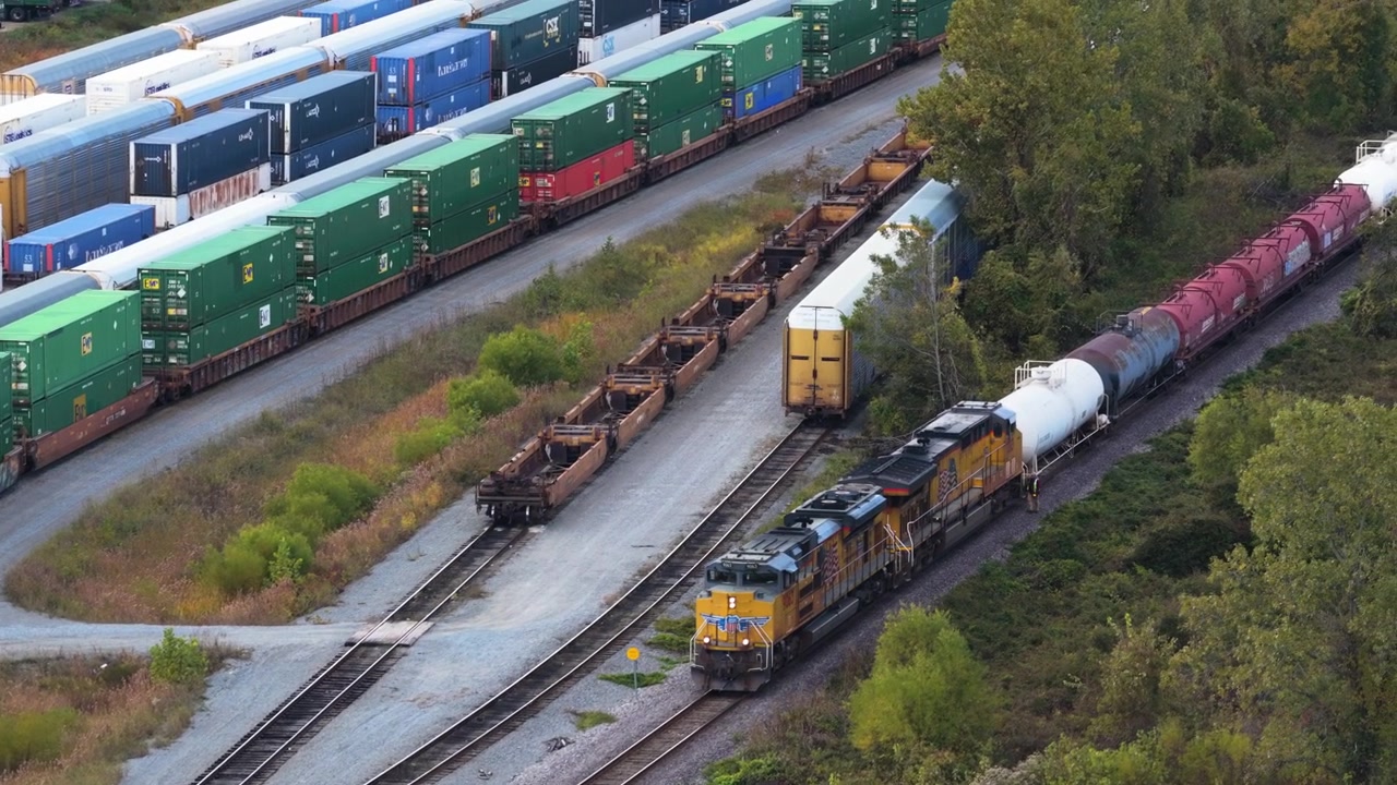 Ripresa aerea ravvicinata di locomotive merci e binari ferroviari accanto a un deposito container, con rivelazione tramite dolly aereo del drone.