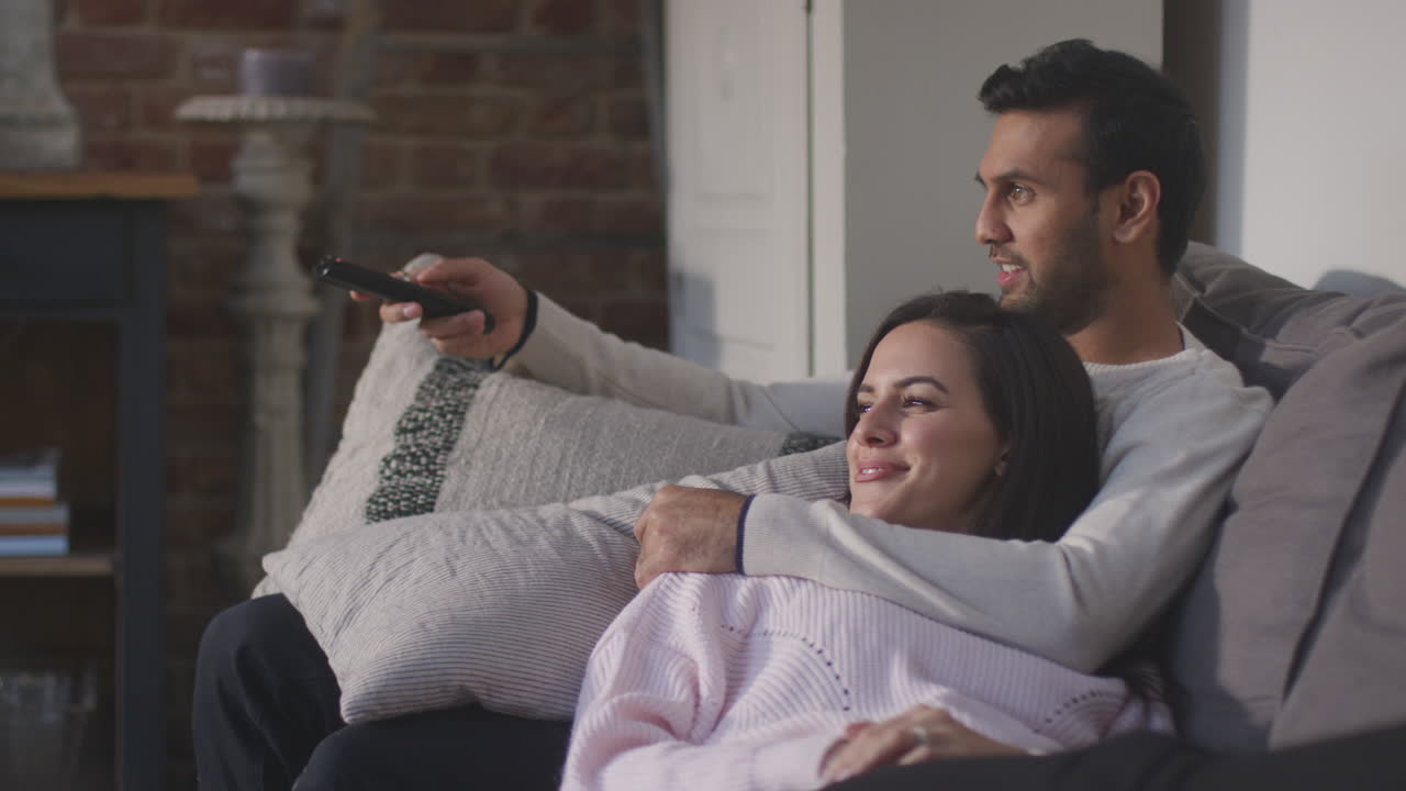 Coppia sdraiata e rilassata sul divano di casa, che guarda la TV insieme.
