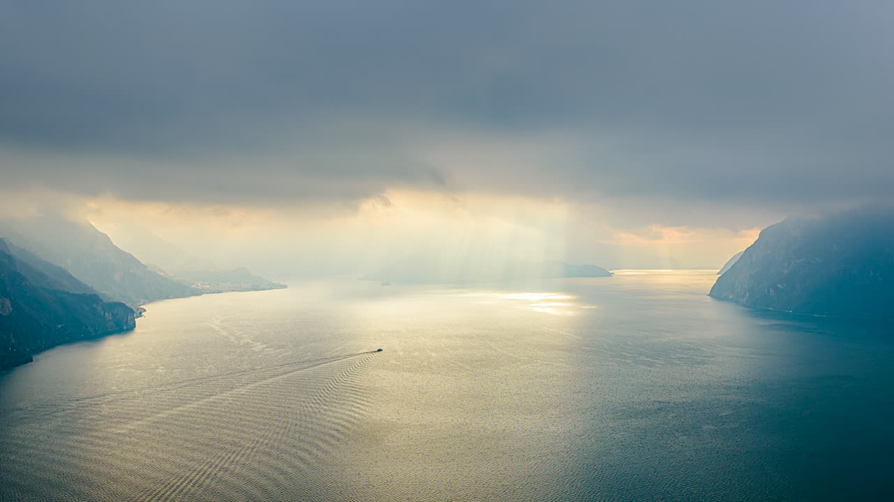Suggestivo timelapse del Lago d'Iseo con nuvole in movimento, soffici raggi di sole e silhouette nebbiose delle montagne che si riflettono sull'acqua calma, creando un suggestivo paesaggio cinematografico italiano