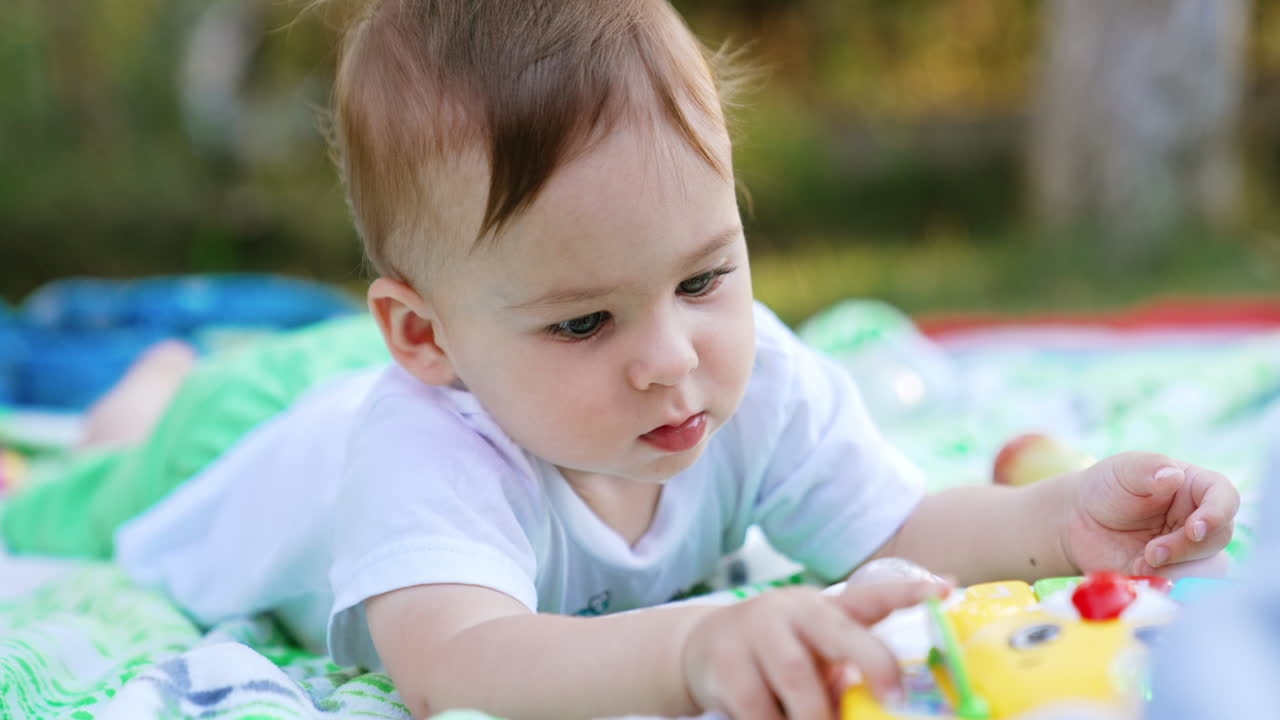 Calm Caucasian kid lies peacefully outdoors. Cute little child busy with his bright toy. Blurred backdrop. Close up.