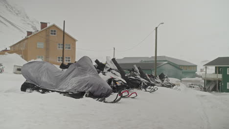 These-snowscooters-are-parking,-with-one-covered-in-a-protective-sheet,-next-to-a-snowy-road-during-heavy-arctic-winds-in-Longyearbyen,-Norway