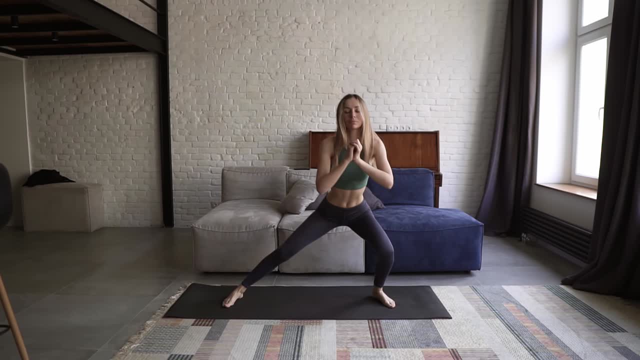 Donna che fa squat laterale su un tappetino da yoga nel salotto di casa con un divano sullo sfondo.