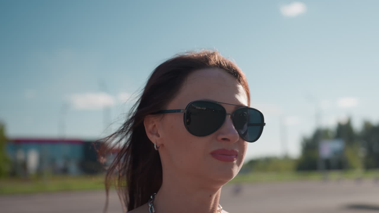 Middle age woman with tattooed hand and chain necklace in leather top outdoors under sunlight, looking confident and stylish with hair moving slightly in wind and blurred urban background