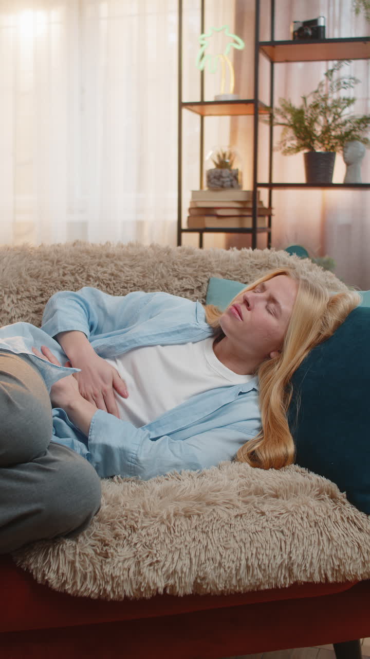 Young woman clutching stomach at home curled posture sofa distress abdominal pain anxious mood