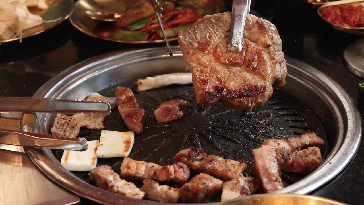 Tongs cut grilled pork belly on tabletop grill, warm lighting, close-up, lively dining atmosphere